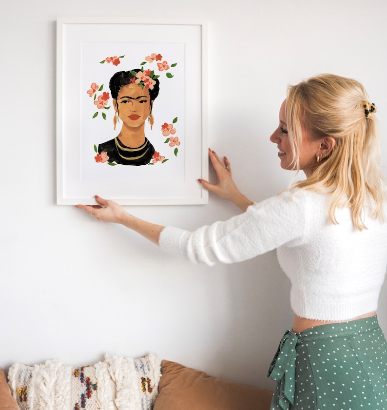 May include: A white framed print of a painting of Frida Kahlo with flowers in her hair. The painting is on a white wall and a person is hanging it up.