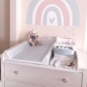 Puede incluir: Una cómoda blanca con un cambiador gris estampado, un conejito de peluche y accesorios para bebés. Los accesorios incluyen una cesta azul y blanca, zapatos de bebé y un chupete. Una calcomanía de arcoíris está encima de la cómoda.