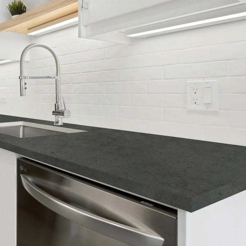 May include: A modern kitchen countertop with a dark gray surface. The countertop is paired with a stainless steel dishwasher and a stainless steel faucet. White subway tile backsplash and white cabinets complete the kitchen design.
