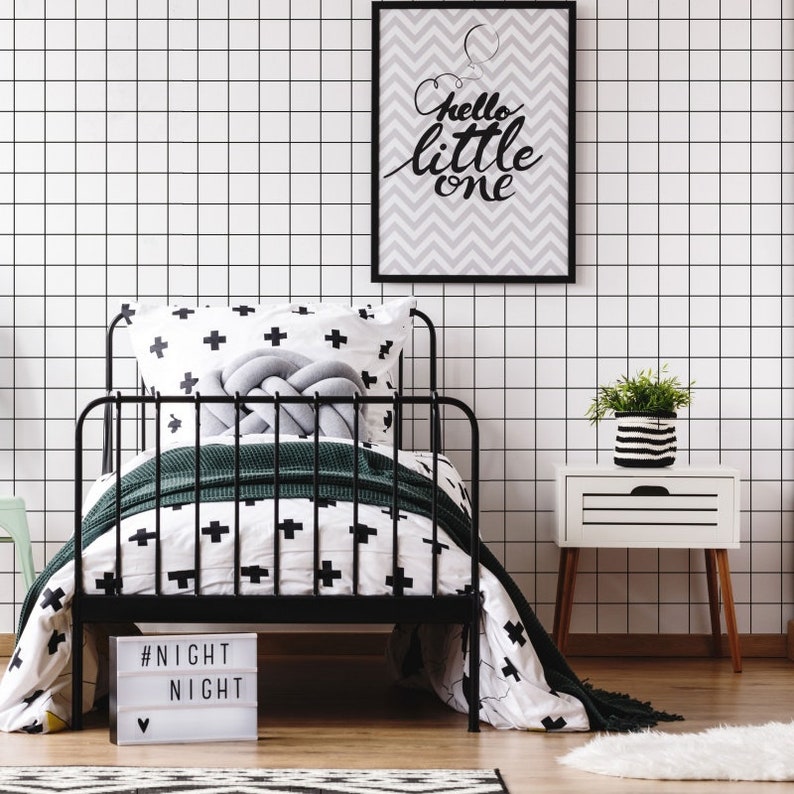 May include: Black metal bed frame with a white and black patterned duvet and pillow. A green throw blanket is draped over the foot of the bed. A white nightlight with the text "#NIGHT NIGHT" is on the floor in front of the bed. A framed print with the text "Hello little one" is hanging on the wall behind the bed. A white nightstand with a black and white striped planter with a green plant is next to the bed.