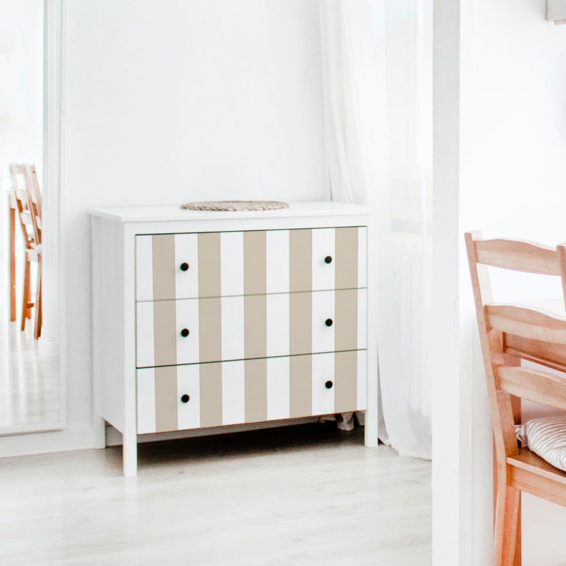 May include: A white chest of drawers with three drawers, each drawer has a beige and white striped pattern. The chest of drawers has black knobs.