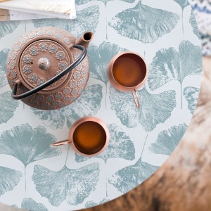 K&ouml;nnte beinhalten: Ein wei&szlig;er Tisch mit einem blauen und wei&szlig;en Ginkgoblatt-Muster. Eine braune Teekanne und zwei Kupferbecher mit Tee stehen auf dem Tisch.