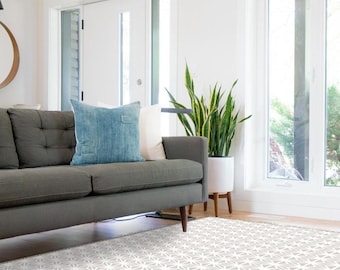 Puede incluir: Un sofá gris con un cojín azul se encuentra sobre una alfombra con estampado geométrico blanco y gris en una sala de estar con una ventana grande y una planta en maceta.
