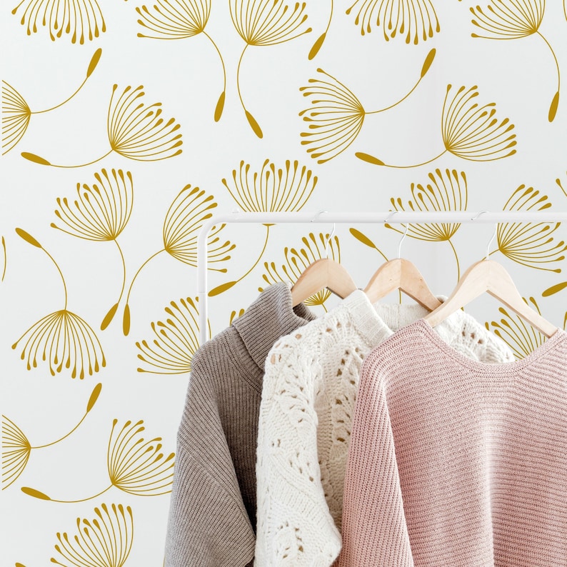 May include: A white wall with a gold dandelion pattern. Three sweaters hang on a white rack against the wall.