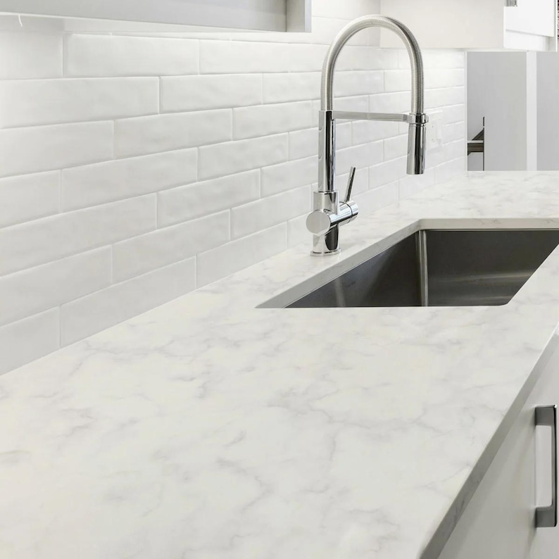 May include: A stainless steel high-arc kitchen faucet positioned over a rectangular sink basin. The countertop features a white marble pattern, and the wall is covered in white rectangular subway tiles with a glossy finish.