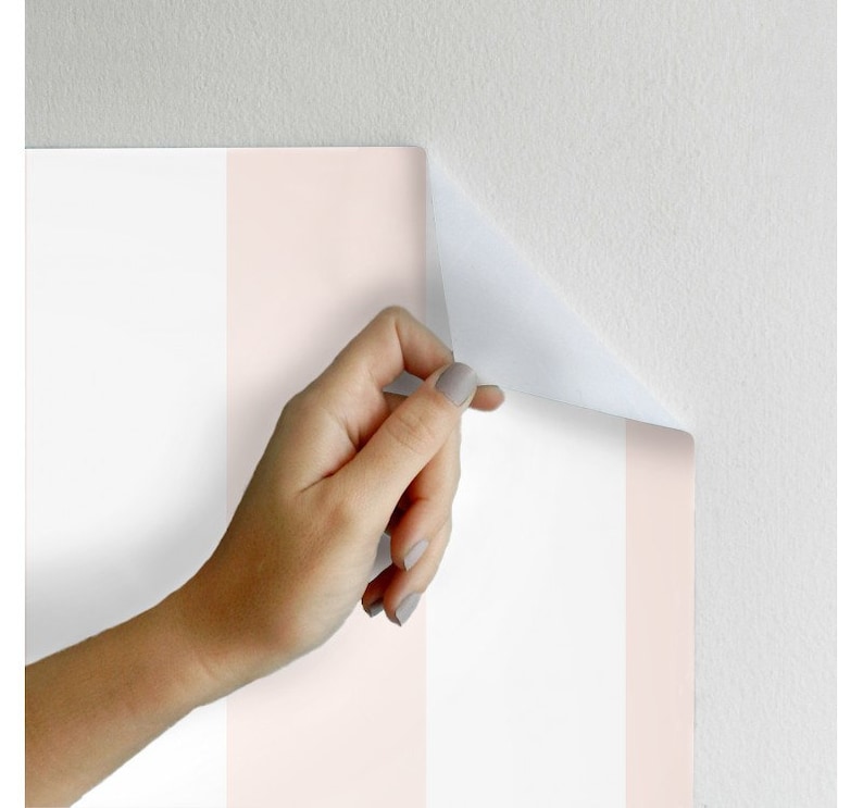 May include: A close-up of pink and white striped peel and stick wallpaper being applied to a wall. The wallpaper is being peeled back to show the adhesive backing.