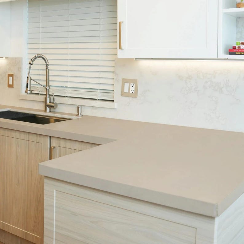 May include: A modern kitchen with light wood cabinets, a gray countertop, and a stainless steel faucet. The cabinets have gold-colored handles. A window with white blinds is visible in the background.