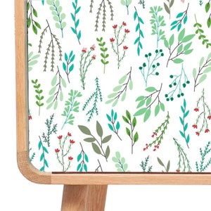 May include: A wooden cabinet with a white surface featuring a repeating pattern of green and blue leaves and red flowers. The cabinet has a light wood finish and four legs.