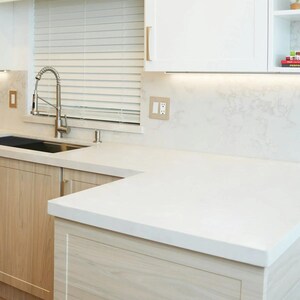 May include: A modern kitchen with light wood cabinets, a white countertop, and a stainless steel faucet. The kitchen features a sink, a window with blinds, and a white backsplash. The overall design is clean and minimalist.