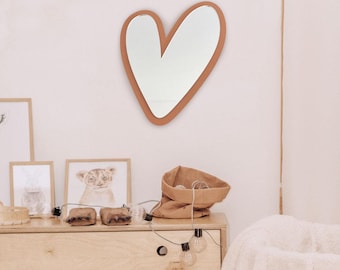 May include: A heart-shaped mirror with a brown frame hangs on a white wall. The mirror is reflecting a room with a dresser, a brown paper bag, and a white blanket.