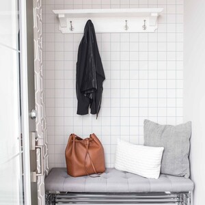 May include: A white shelf with hooks hangs above a gray bench with a brown leather bag and two pillows. The wall behind the shelf is tiled with white square tiles.