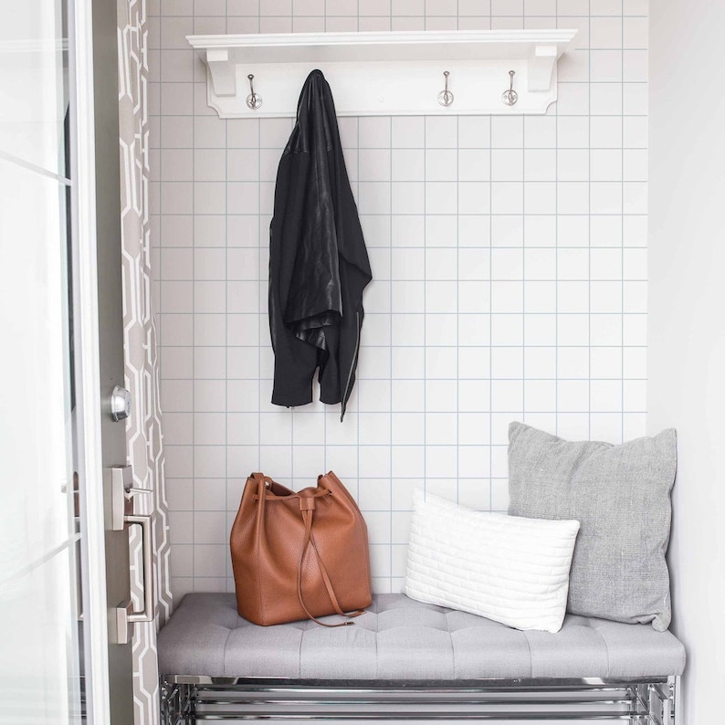 May include: A white shelf with hooks hangs above a gray bench with a brown leather bag and two pillows. The wall behind the shelf is tiled with white square tiles.