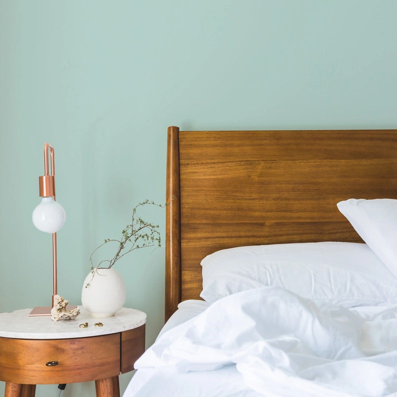 Op de afbeelding: Een wit bed met een houten hoofdeinde en witte beddengoed. Een klein nachtkastje met een lamp en een vaas met groen staat naast het bed.