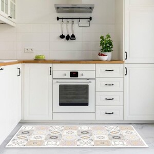Op de afbeelding: Een witte keuken met witte kasten en een zwarte oven. Voor de kasten ligt een gepatroneerd keukenkleed met beige, grijze en witte tegels.