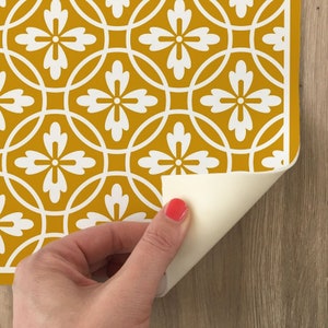May include: A sheet of peel and stick wallpaper with a repeating pattern of white flowers on a mustard yellow background. The wallpaper is being peeled back to reveal the adhesive backing.