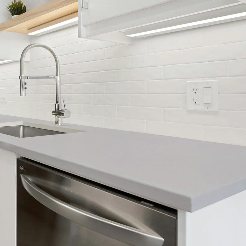 May include: A modern kitchen with a stainless steel dishwasher, a stainless steel faucet, and a gray countertop. The backsplash is white brick, and there is a small plant on a wooden shelf above the sink.