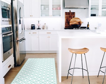 Puede incluir: Una cocina blanca con una nevera de acero inoxidable, armarios blancos y una isla blanca con dos taburetes de bar de madera. Una alfombra de color azul claro y blanco con un patrón está en el suelo.