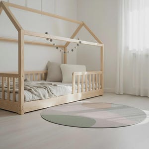 May include: A light wood-framed bed with a house-shaped top, featuring a mattress, pillows, and a blanket. A round rug with abstract shapes in muted green, pink, and peach tones sits on the floor. A garland of gray felt balls hangs above the bed.