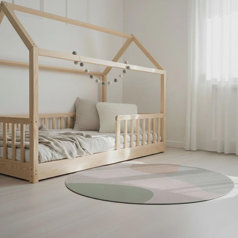 May include: A light wood-framed bed with a house-shaped top, featuring a mattress, pillows, and a blanket. A round rug with abstract shapes in muted green, pink, and peach tones sits on the floor. A garland of gray felt balls hangs above the bed.