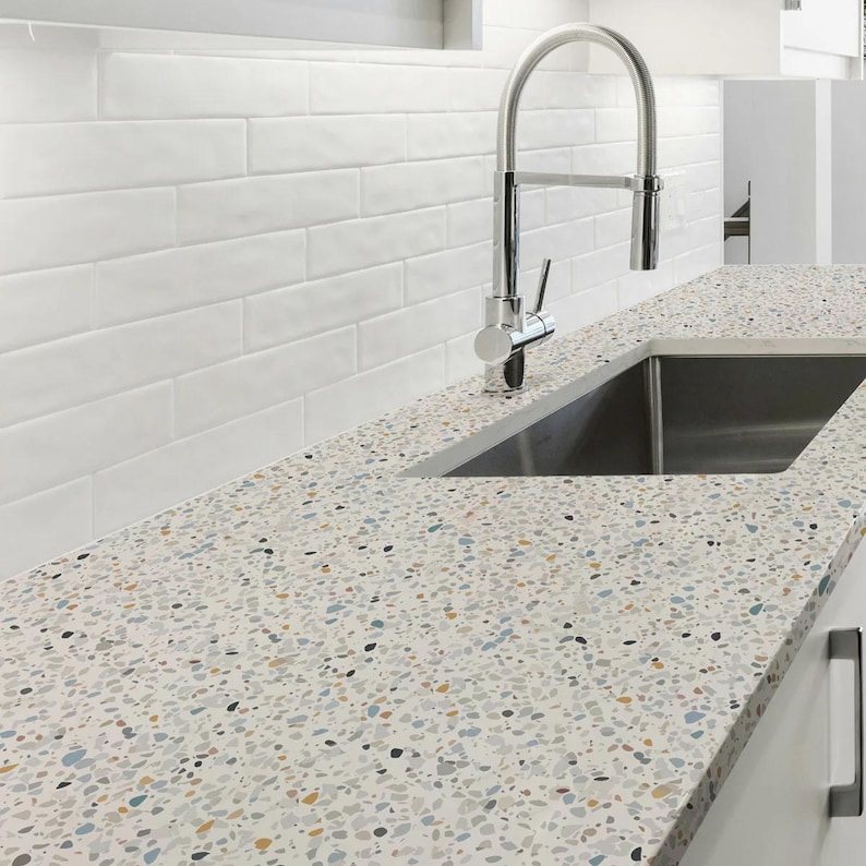 May include: A kitchen countertop with a speckled pattern in shades of white, gray, blue, and gold. A stainless steel sink and faucet are installed. White subway tile backsplash.