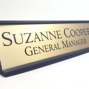 May include: Gold desk nameplate with black text that reads "Suzanne Cooper General Manager".