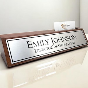 May include: A wooden desk nameplate with a silver nameplate that reads "Emily Johnson, Director of Operations." A business card is in a slot on top. The nameplate is on a white surface.