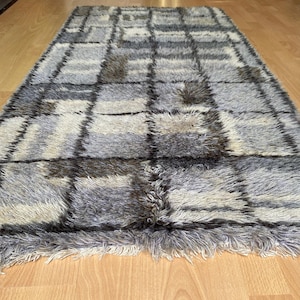 May include: A rectangular rug with a geometric pattern. The rug features a grid design with alternating squares of gray, beige, and dark brown. The rug has a shaggy texture and a fringe along the bottom edge.