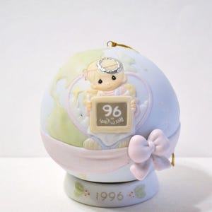 May include: A ceramic ornament featuring a cherubic figure holding a sign with "96" and "Precious Gem". The ornament is globe-shaped, with a light blue and green color scheme, a pink ribbon, and a bow. The base reads "1996".