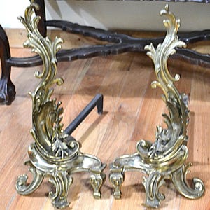 May include: Pair of antique brass andirons with ornate, swirling designs. The andirons have a decorative, curved shape with a black metal bar connecting them. The brass has a warm, aged patina, adding to their vintage appeal. These are likely used for a fireplace.