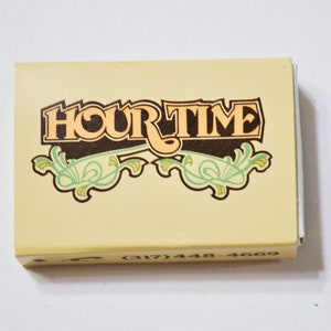 May include: A yellow matchbox with the text "HOUR TIME" in brown and orange lettering with a green and brown floral design. The phone number (317) 448-4669 is printed on the bottom of the box.