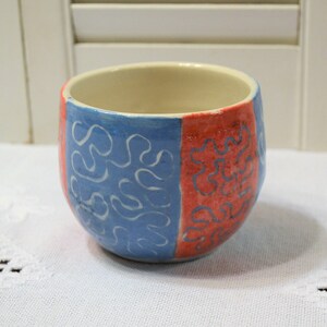 May include: A handmade ceramic cup with a rounded shape. The cup is divided into two vertical sections, one blue with white squiggly lines and the other red with similar white lines. The interior is a light beige color.