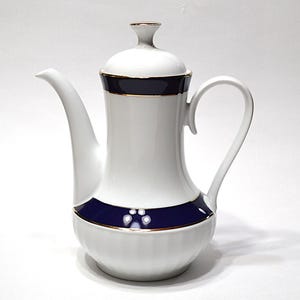 May include: White ceramic coffee pot with a navy blue band and gold trim. The pot has a curved spout and a handle. The lid is white with a navy blue band and a small gold trim. The coffee pot is on a white background.