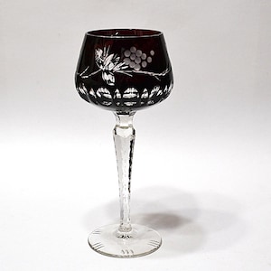 May include: A burgundy wine glass with a clear, etched stem and base. The bowl has an etched design of grapes and leaves. The glass is designed for wine and is set against a white background.