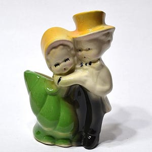May include: A vintage ceramic figurine of a couple embracing, with the woman wearing a yellow bonnet and the man in a yellow top hat. The couple is hugging a green leaf-shaped vase. The figurine is painted in soft, muted colors.