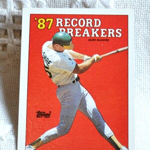 May include: A baseball card featuring a baseball player in a green and white uniform swinging a bat. The card is red with white lettering that reads '87 Record Breakers' and 'Mark McGwire'. The card is from Topps.