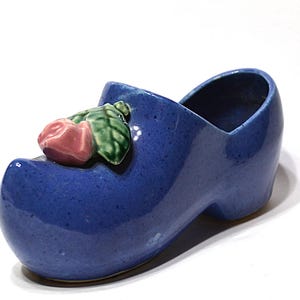 May include: A blue ceramic clog-shaped planter with a pink rose and green leaves on the front. The planter is a vibrant blue color and has a glossy finish. The clog is a decorative item.