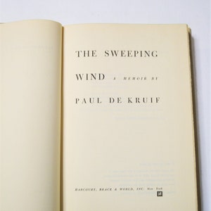 May include: Open book with the title "THE SWEEPING WIND" by Paul De Kruif. The book's pages are off-white, and the text is in black. The publisher's name, Harcourt, Brace & World, Inc., is at the bottom.