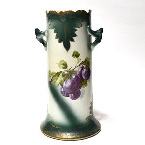May include: A cylindrical porcelain vase featuring a cream-colored body with dark green gradients at the top and bottom. The vase is decorated with a painted cluster of purple grapes and green leaves, accented by gold trim along the scalloped rim.