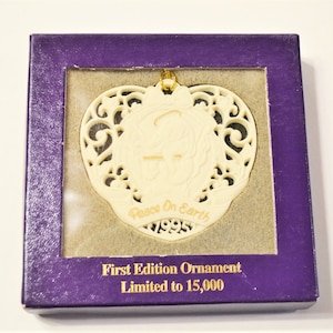 May include: Ivory-colored heart-shaped ornament with intricate cut-out design, featuring the words "Peace On Earth" and the year "1995." The ornament is in a purple box with the text "First Edition Ornament" and "Limited to 15,000."