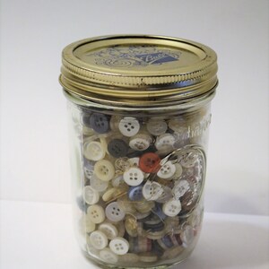 May include: A clear glass jar filled with a variety of buttons in different colors and sizes. The jar has a gold-colored lid and is labeled with the word "Ball". The buttons include white, beige, gray, blue, and red.