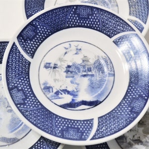May include: A set of white ceramic plates with blue decorative patterns. Each plate has a central landscape scene in blue and white, surrounded by a patterned border. The plates are arranged in a circular pattern.