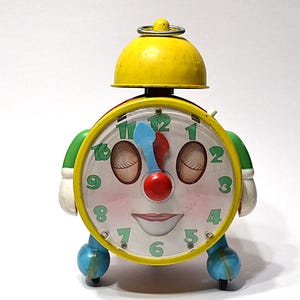 May include: A vintage-style alarm clock with a yellow bell on top and a round face featuring a cartoon character design. The clock has green numbers, a blue hour hand, a red minute hand, white gloved hands on the sides, and blue feet.