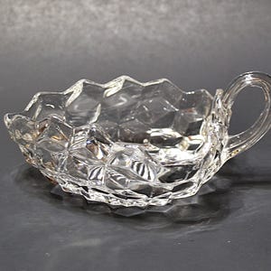 May include: Clear glass serving dish with a handle. The dish has a diamond-patterned design and a scalloped rim. The handle is a loop shape. The dish is sitting on a dark surface.