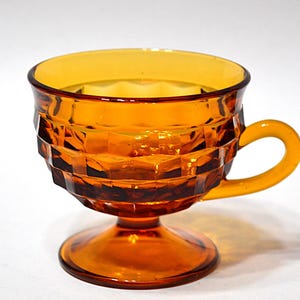 May include: Amber-colored glass teacup with a geometric pattern. The cup has a rounded bowl, a small handle, and a pedestal base. The glass is transparent, allowing light to pass through, and is set against a white background.