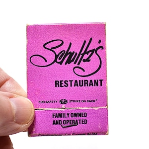 May include: A rectangular pink matchbook with black text that reads Schultz's RESTAURANT, FOR SAFETY STRIKE ON BACK, and FAMILY OWNED AND OPERATED. The matchbook features a script logo and is printed on a textured paper surface with visible wear.