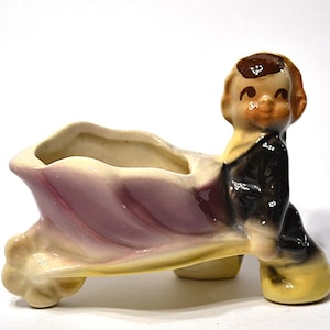 May include: A vintage ceramic planter in the shape of a boy pushing a wheelbarrow. The planter features a boy with brown hair and a black suit. The wheelbarrow is light purple and cream-colored.