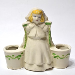 May include: A vintage ceramic figurine of a girl in a white dress and bonnet, holding two small buckets. The figurine has blonde hair, blue eyes, and green accents on the buckets and ropes.