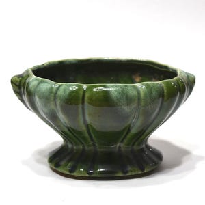 May include: A dark green ceramic bowl with a scalloped, fluted edge and a pedestal base. The glaze features a glossy finish with lighter green highlights along the ridges of the vertical fluting, creating a textured appearance on the exterior.