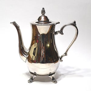 May include: A silver-colored coffee pot with a curved spout and handle. The pot has a lid with a decorative finial and rests on three ornate feet. The polished surface reflects the surrounding environment.