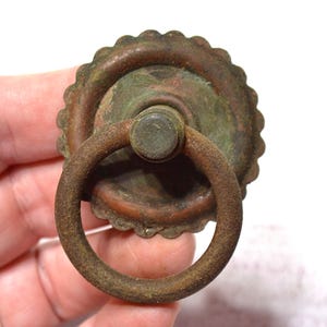 Peut inclure: Une poignée de tiroir circulaire en métal avec une finition brune rouillée et patinée. La quincaillerie présente une plaque arrière à bord festonné et une poignée en anneau articulée fixée sur un montant central, montrant des signes d'oxydation.